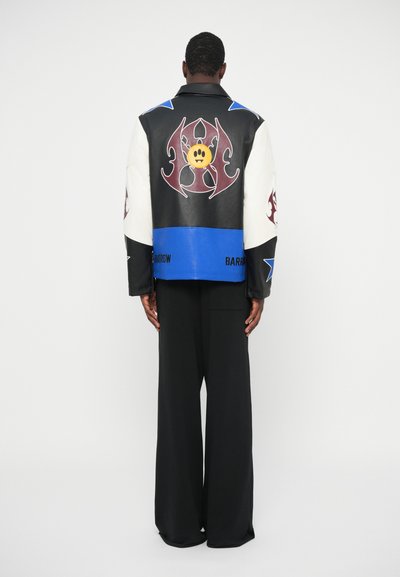 Schwarze und weiße Lederjacke mit bunten Grafiken, darunter ein gelbes Gesicht und stilisierte Designs, mit lockerem Schnitt und weiten Ärmeln.