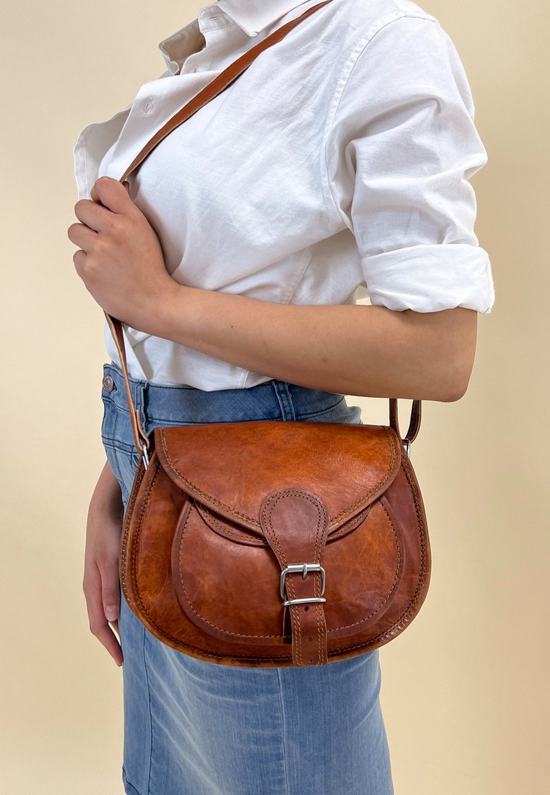 Braune Leder-Crossbody-Tasche mit einem geschwungenen Deckel, silbernem Schnallen-Detail und verstellbarem Trageriemen. Die Oberfläche ist glatt mit minimaler Naht.