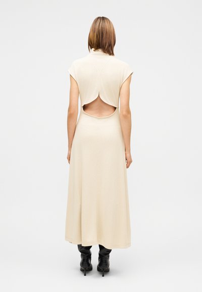 Μπεζ φόρεμα με κοντά μανίκια, ανοιχτή πλάτη και ροής φούστα. Λεία υφή, μήκος μέχρι το γόνατο, φορεμένο με μαύρες μπότες με τακούνι.
