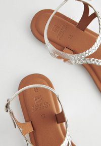 Sandali piatti marroni con cinturini intrecciati in argento metallico. La soletta in pelle sintetica testurizzata presenta il marchio e i dettagli della taglia.