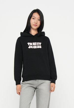 Svart hettegenser laget av mykt stoff med en lys rosa "TOMMY JEANS"-logo i en rund, leken skrifttype. Elastiske mansjetter og linning.