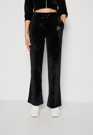 Person trägt schwarze Samt-Hose mit weitem Bein und Kordelzug in der Taille sowie kleinem Strassdesign auf der linken Tasche, kombiniert mit weißen Sneakern.