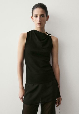 Donna con capelli scuri che indossa un top nero senza maniche con un dettaglio ad anello su una spalla, abbinato a una gonna nera e collant neri trasparenti.