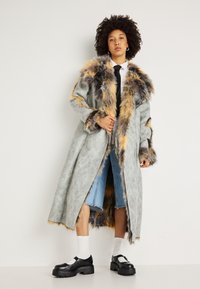 Garšs pelēks mākslīgās kažokādas mētelis ar daudzkrāsainām akcentiem, apvienots ar baltu krekli, melnu kaklasaiti, zilu džinsu šortiem un masīvām melnām kurpēm.