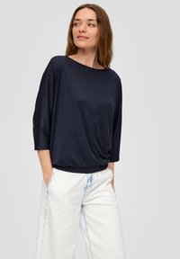Donkerblauwe, lichte top met een losse pasvorm, brede halslijn en driekwartmouwen, gecombineerd met witte broek met lichte blauwe slijtage.