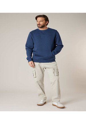 Man wearing navy blue sweatshirt, beige cargo pants, and white sneakers standing with one hand in pocket and looking left against plain background.