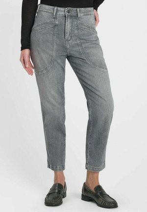 Femme portant un jean gris taille haute avec des détails de coutures avant courbées et des mocassins noirs, debout devant un fond uni.