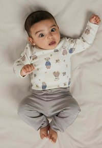 Spädbarn som ligger på lätt tyg, iklädd en vit långärmad skjorta med teddy-björntryck och grå byxor, tittar uppåt med en öppen mun.