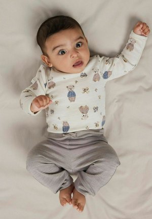 Bébé allongé sur un tissu léger, portant une chemise blanche à manches longues avec des motifs de nounours et un pantalon gris, regardant vers le haut avec la bouche ouverte.