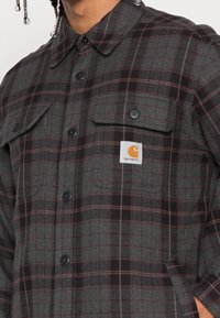 Rutet flannelskjorte i grå, sort og burgunder. Har to brystlommer, sorte knapper og et Carhartt-logo patch på forsiden.