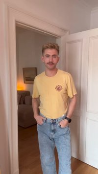 Joven con cabello corto rubio y bigote, vistiendo una camiseta amarilla de Element y jeans azules, de pie en una puerta con las manos en los bolsillos.