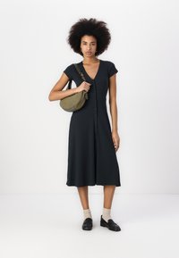 Schwarzes Hemdkleid mit V-Ausschnitt, kurzen Ärmeln und schwingendem Rock. Das Model hält eine olivgrüne Umhängetasche; kombiniert mit hellen Socken und Slippern.