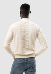 Uomo visto da dietro che indossa un maglione crema con testo verticale "BESS BI KK" e jeans blu su uno sfondo grigio uniforme.