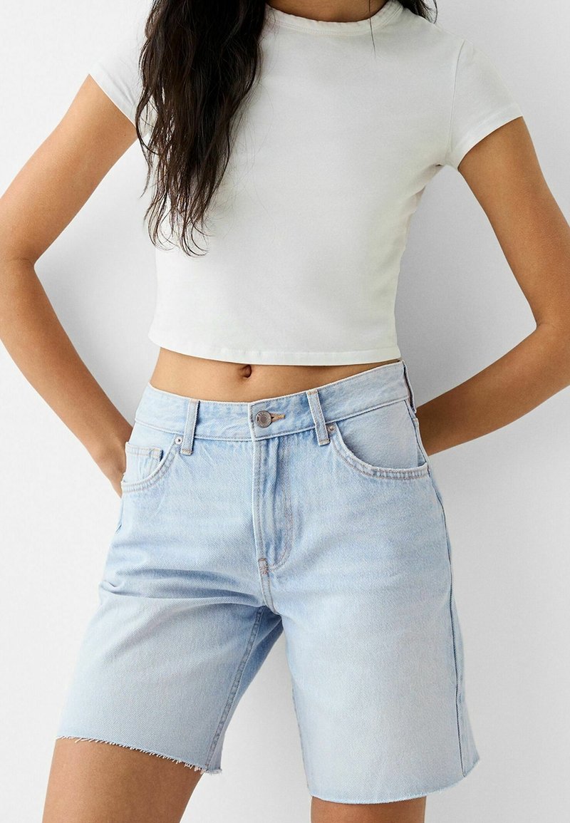 Kvinna i en vit kortärmad t-shirt och ljusblå högmidjade denimshorts med slitna kanter, stående med händerna i fickorna.