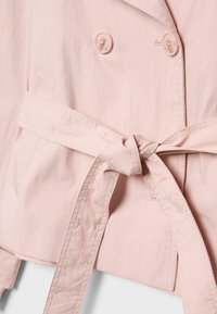 Světle růžová bunda s dvojitým řaděním knoflíků a látkovým páskem zavázaným v pase, s detailním prošíváním a knoflíkovými dírkami.