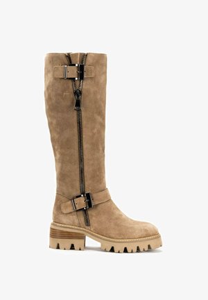 Bota de ante color beige hasta la rodilla con cremallera lateral, detalle de hebilla y tacón grueso texturizado. Presenta una suela robusta con surcos pronunciados.
