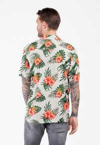 Mann mit Tattoos, der ein kurzärmeliges weißes Hemd mit roten Hibiskusblüten und grünen Blättern trägt, steht mit dem Rücken zu einem weißen Hintergrund.