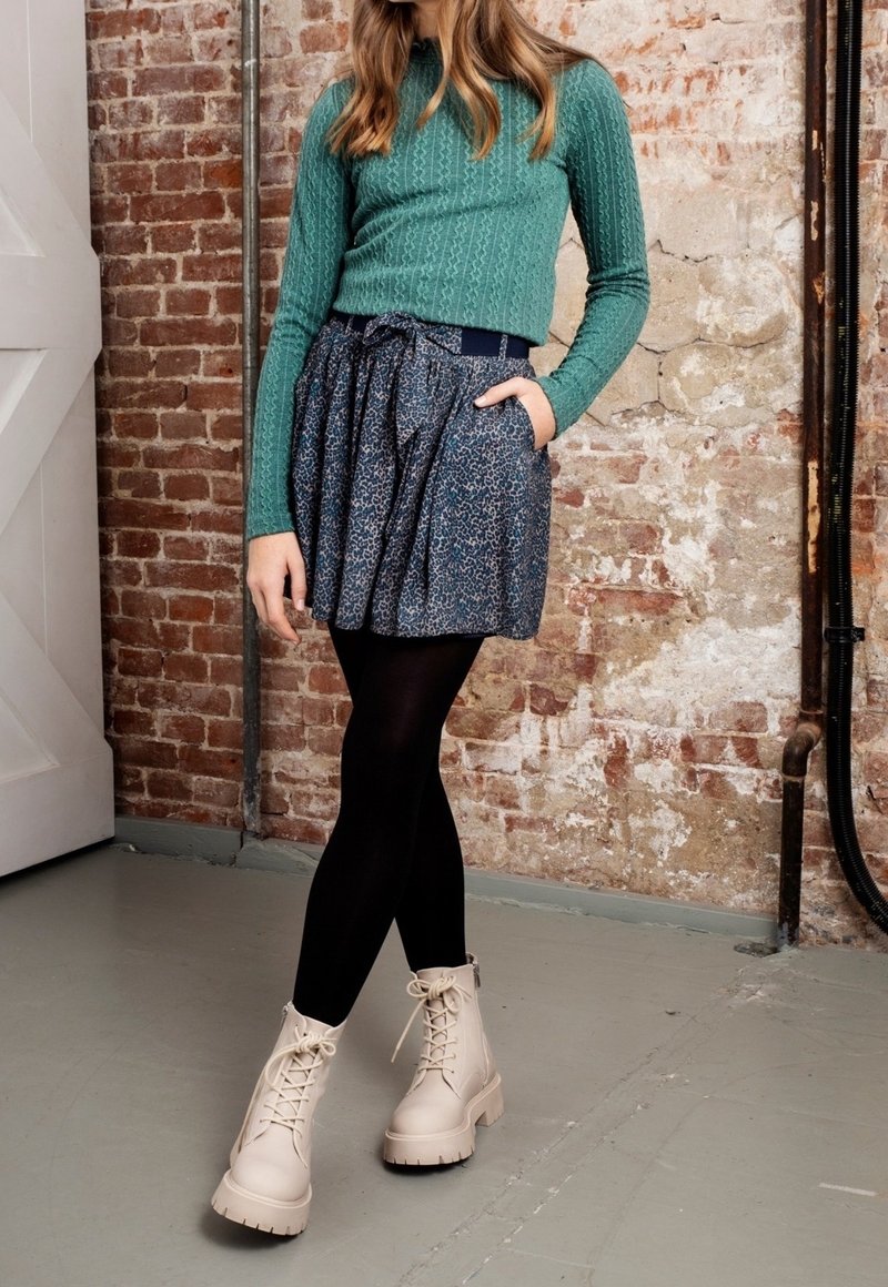 Zelený žebrovaný svetr s dlouhým rukávem spárovaný s námořnickou sukní s vzorem, nošený s černými punčochami a krémovými šněrovacími kotníkovými botami. Texturované a vrstvené.