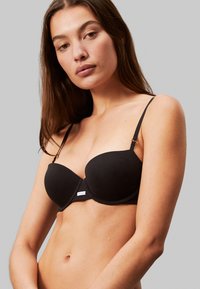 Reggiseno nero senza spalline realizzato in tessuto liscio, con coppe sagomate, spalline regolabili e un piccolo logo al centro davanti.