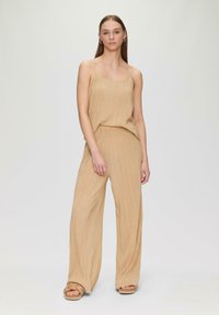 Beige geplooide jumpsuit met dunne bandjes, wijde benen en een relaxte pasvorm. Soepele textuur met subtiele glans, gecombineerd met chunky sandalen.