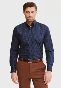 Granatowa koszula z długim rękawem w mały wzór w kropki, z guzikami z przodu i kołnierzykiem. Noszona z brązowymi spodniami i brązowym paskiem.