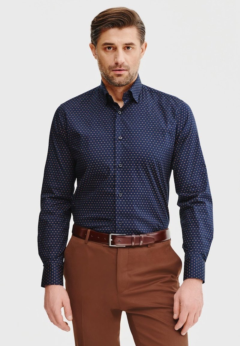 Granatowa koszula z długim rękawem w mały wzór w kropki, z guzikami z przodu i kołnierzykiem. Noszona z brązowymi spodniami i brązowym paskiem.