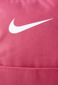Ružová batoh Nike s textúrovanou tkaninou a bielym logom. Dizajn obsahuje horné sťahovacie uzatváranie a plochý detail vrecka.