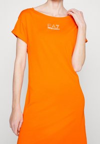 Kvinna som bär en klar orange klänning med korta ärmar och EA7 Emporio Armani-logotypen på bröstet, hand uppsträckt nära axeln.