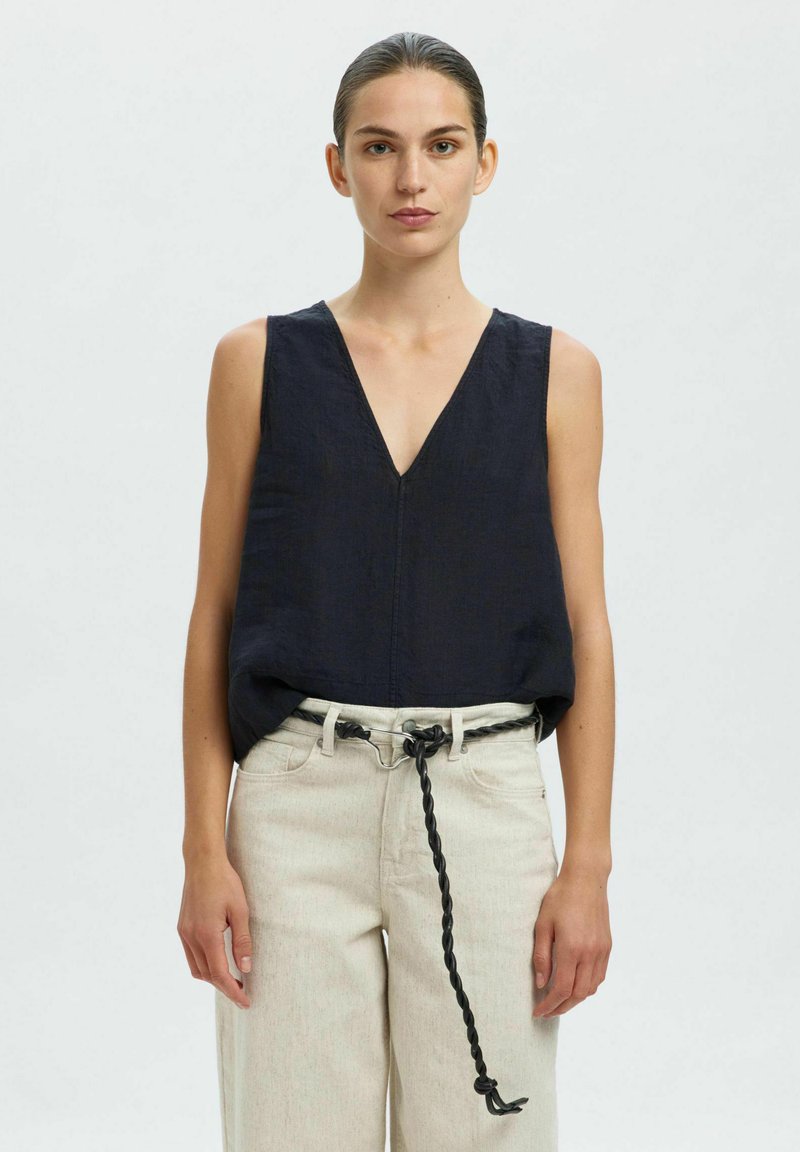 Haut sans manches en lin bleu marine avec un décolleté en V prononcé, associé à un pantalon large beige clair et un détail de ceinture tressée noire.
