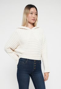 Kremkrāsas cropped kapuci ar ribotā tekstūra, kurai ir priekšējā kabata un kapuce ar auklu, apvienots ar tumši ziliem skinny džinsiem.