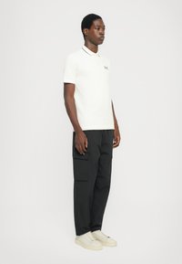 Polo shirt in off-white met zwarte kraag en geborduurde logo, gecombineerd met losse zwarte cargobroeken en witte sneakers.