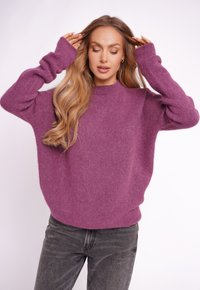 Oversized, long-sleeve sweater in soft, fuzzy purple material. Features a high neckline and ribbed cuffs, paired with fitted gray jeans.