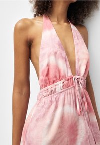 Mujer vestida con un vestido sin mangas de halter en colores rosa y blanco con un patrón de tie-dye, con un escote en V profundo y cintura ceñida, con cabello rizado visible contra un fondo blanco.