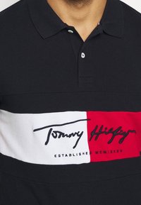 Camisa polo negra con una franja horizontal blanca y roja, que cuenta con el logo de Tommy Hilfiger bordado en negro. Cuello clásico y botones visibles.