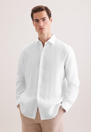Homme portant une chemise blanche à manches longues boutonnée et un pantalon beige, les mains dans les poches, sur un fond clair uni.