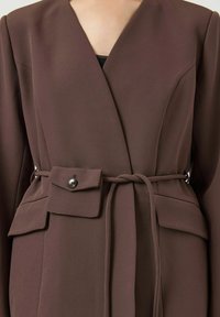 Bruine wikkeljurk met een diepe V-hals, lange mouwen, een bijpassende riem en een zijzak met knoopdetail. Gladde stof met een gestructureerd ontwerp.