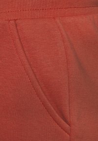 Punainen tekstuurinen kangas, jossa on näkyvä tasku ja ribattu vyötärö. Materiaali näyttää venyvää ja pehmeää, sopivaa rentoon pukeutumiseen.