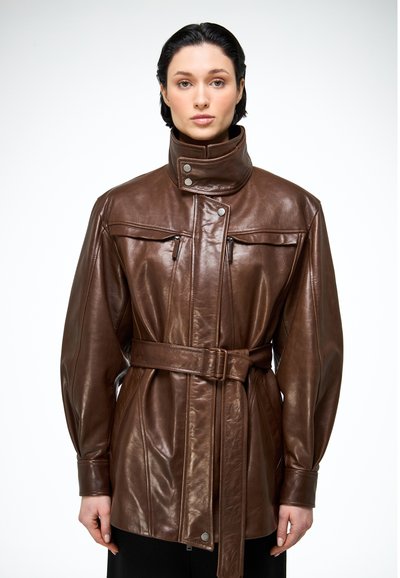 Femme portant une veste en cuir marron ceinturée avec un col montant et des poches zippées sur la poitrine, debout devant un fond blanc uni.