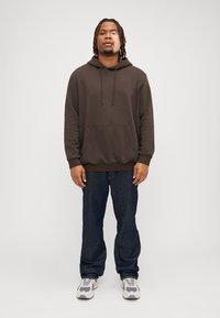 Brun hettegenser med kengurulomme, kombinert med mørkeblå jeans og lysegrå sneakers. Stoffet ser mykt og teksturert ut.