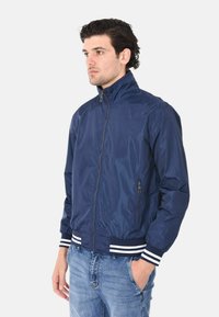 Giacca bomber in nylon blu navy con colletto alto, chiusura frontale a zip, tasche laterali e righe bianche sui polsini e sull'orlo.