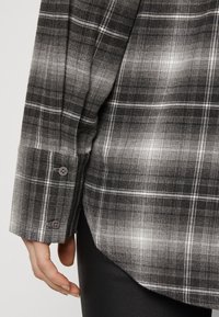 Camicia plaid grigia con motivo a quadri neri e bianchi, con polsini a bottone e orlo leggermente curvo. Realizzata in un tessuto morbido e testurizzato.
