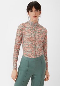 Transparente Bluse mit hohem Kragen, ausgestattet mit einem mehrfarbigen, abstrakten Muster aus Koralle, Grün und Beige; kombiniert mit hochgeschnittenen tealfarbenen Hosen.