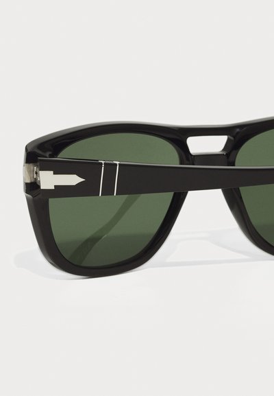 Occhiali da sole neri con montatura spessa, lenti verdi e dettagli in metallo argentato con un design a freccia sulle tempie. Texture liscia e lucida.