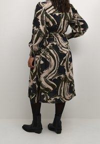 Robe longue noire avec un motif abstrait beige et vert, taille cintrée, manches amples et ourlet à la cheville, associée à des bottines noires.