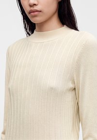 Jeune femme portant un pull en maille côtelée de couleur crème avec un col montant et des manches longues, visible de la poitrine jusqu'au menton.