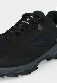 Μαύρο πεζοπορικό παπούτσι Columbia με υφασμάτινες υφές και πλέγμα, κορδόνια με σχέδιο, χοντρή μαλακή σόλα και ασφαλή αγκράφα σε γκρι φόντο.