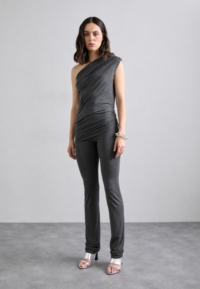 Top grigio asimmetrico a spalla unica con texture arricciata, abbinato a pantaloni grigi aderenti. La modella indossa tacchi a spillo argentati. Finishing in tessuto liscio.