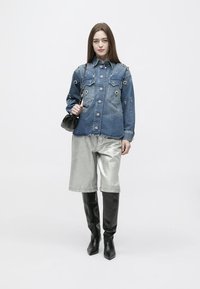 Νεαρή γυναίκα που φορά διακοσμημένο τζιν μπουφάν, μεταλλικά ασημί σορτς, μαύρες μπότες μέχρι το γόνατο και κρατά μια μικρή μαύρη τσάντα.