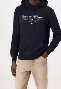 Felpa blu navy con tasca frontale e lacci regolabili. Presenta testo bianco e rosso: "Tommy Hilfiger New York City Dal 1985."