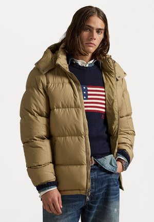 Chaqueta de plumas beige con capucha, diseño acolchado, cierre frontal con cremallera y bolsillos laterales. Llevada sobre un suéter azul marino y jeans.